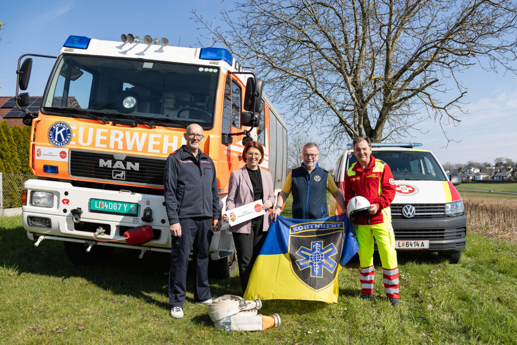 Margot Haag zeigt eine Begegnung vor einem orangefarbenen Feuerwehrfahrzeug RLFA 2000; im Gespräch mit Erich Hofmarcher, Landesrätin Langer-Weninger und einem weiteren Helfer, farblich auffällig Blau und Gelb im Outfit.